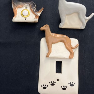 May include: Three ceramic dog-themed light switch covers and a doorbell button. One switch cover features a brown and tan dog, another a gray dog, and the third a brown dog. The doorbell button has a black, brown, and tan dog design.