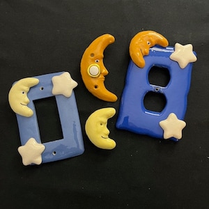 May include: A set of five ceramic light switch covers featuring a moon and star theme. The covers are blue with yellow and white accents. The moon and star shapes are three-dimensional.