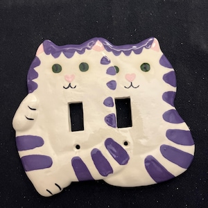 May include: A white ceramic light switch cover featuring two cat faces. The cats have purple stripes and accents, green eyes, and pink heart-shaped noses. The cover has two rectangular openings for the switches.