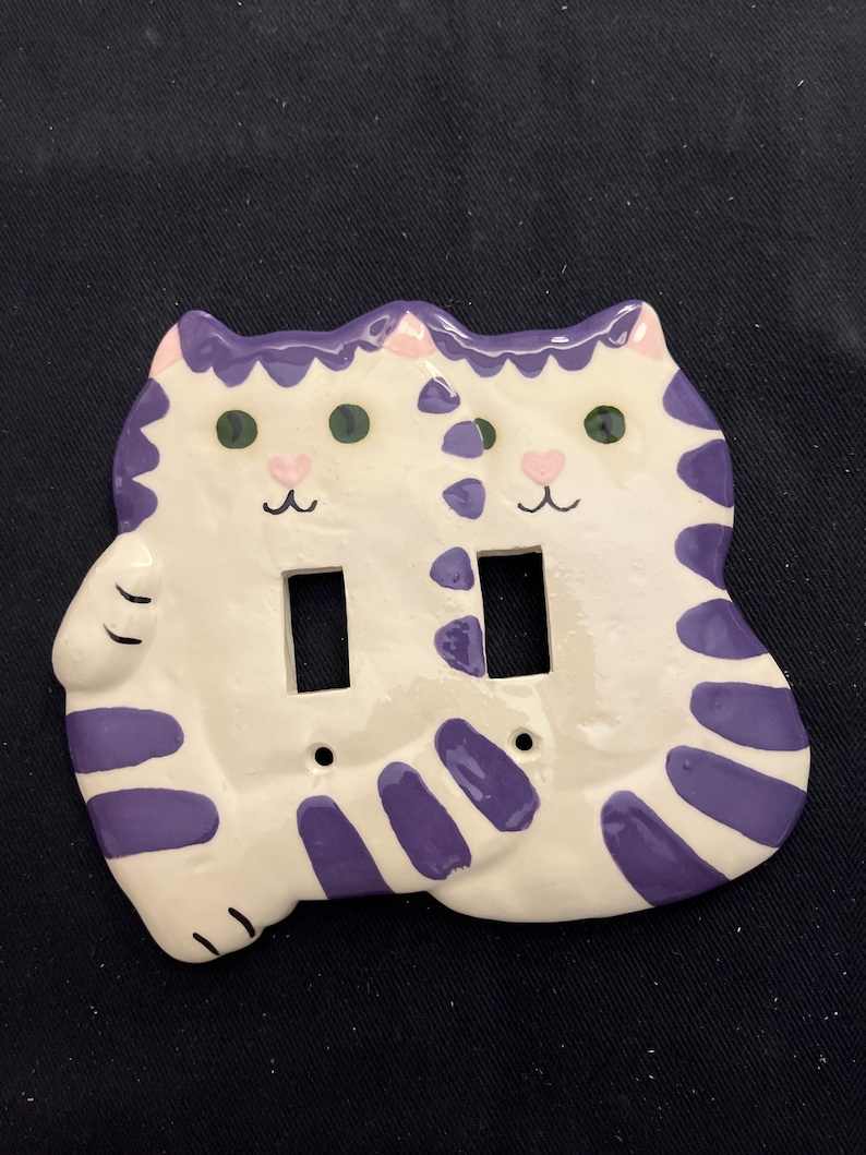May include: A white ceramic light switch cover featuring two cat faces. The cats have purple stripes and accents, green eyes, and pink heart-shaped noses. The cover has two rectangular openings for the switches.