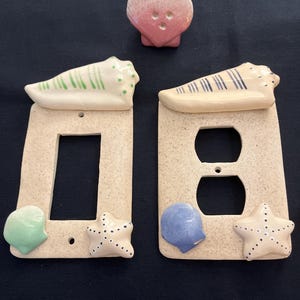 May include: Two ceramic light switch and outlet covers with a beach theme. The covers are beige with a sandy texture. Each cover has a seashell, a starfish, and a small seashell in the corner. A separate ceramic seashell with a pink and white ombre design is also included.