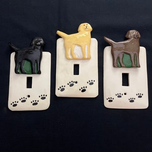 May include: Three ceramic light switch covers featuring a black, yellow, and brown dog silhouette. Each cover has a white background with black paw prints.