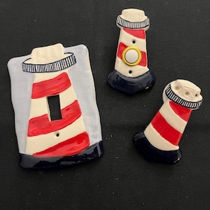 May include: Three ceramic light switch covers shaped like lighthouses with red and white stripes and a blue base. The center cover has a gold ring and a white button.