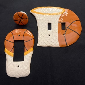 May include: A set of ceramic light switch covers and a knob, featuring a basketball theme. The covers are painted with orange and black basketball designs, and the knob is a basketball shape. The background is black.