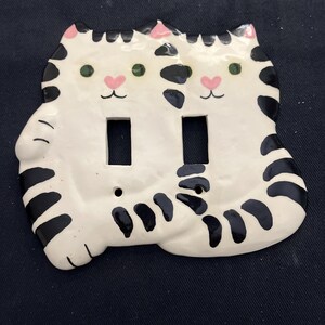 May include: White ceramic double light switch cover featuring two cat faces. Each cat has green eyes, pink nose, and black stripes. The cover has two rectangular openings for the light switches.