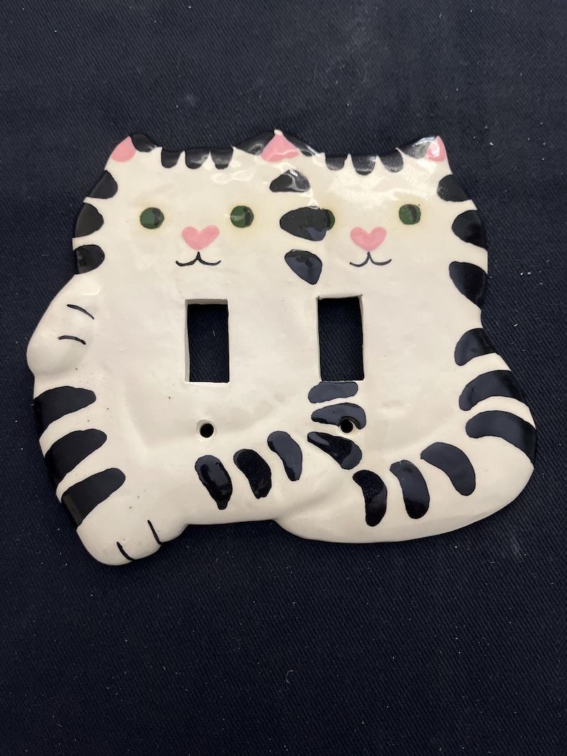 May include: White ceramic double light switch cover featuring two cat faces. Each cat has green eyes, pink nose, and black stripes. The cover has two rectangular openings for the light switches.