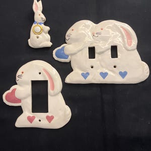 May include: Three ceramic wall plates featuring white rabbit designs. One single-switch plate shows a rabbit with pink hearts, another double-switch plate shows two rabbits with blue hearts, and a small rabbit-shaped wall ornament with a gold ring.