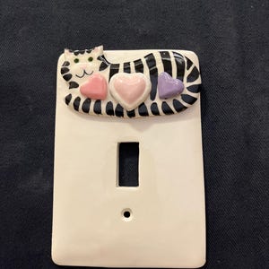 May include: White ceramic light switch cover with a decorative cat design. The cat has black and white stripes, green eyes, and is adorned with three heart-shaped accents in pink and purple.