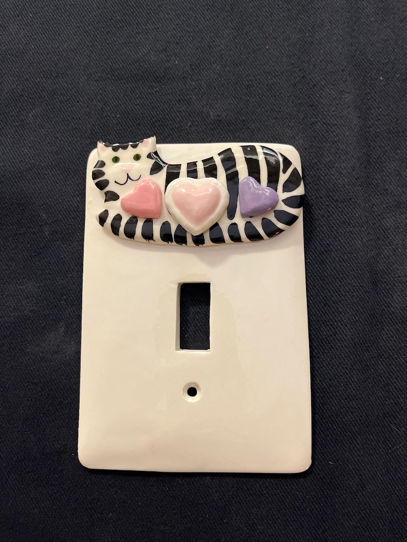 May include: White ceramic light switch cover with a decorative cat design. The cat has black and white stripes, green eyes, and is adorned with three heart-shaped accents in pink and purple.