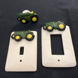 May include: Three white ceramic light switch covers with a green tractor design. The tractor has yellow wheels and a black driver. The switch covers are for a single light switch.
