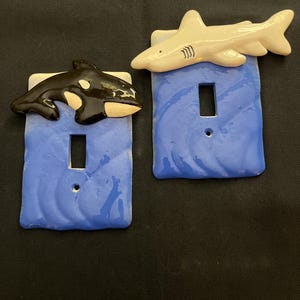 May include: Two ceramic light switch covers with blue backgrounds and ocean-themed designs. One features a white shark and the other a black and white orca.