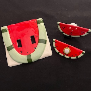 May include: A set of three ceramic watermelon-themed light switch covers. The main cover features a red watermelon with green stripes and two white light switch openings. The two smaller covers are watermelon slices with black seeds and green stripes.