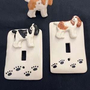 May include: Two white ceramic light switch covers with a brown and white dog illustration and paw prints. A separate brown and white dog figurine.