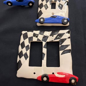 May include: Three ceramic light switch covers with a checkered racing flag design. The top cover features a blue race car, the bottom cover features a red race car, and the middle cover has two blank switch plates.