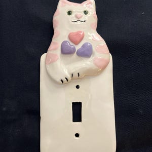 May include: A white ceramic light switch cover featuring a cat design. The cat has pink spots and holds three hearts, one pink and two purple. The switch plate is rectangular with a single switch opening and two screw holes.