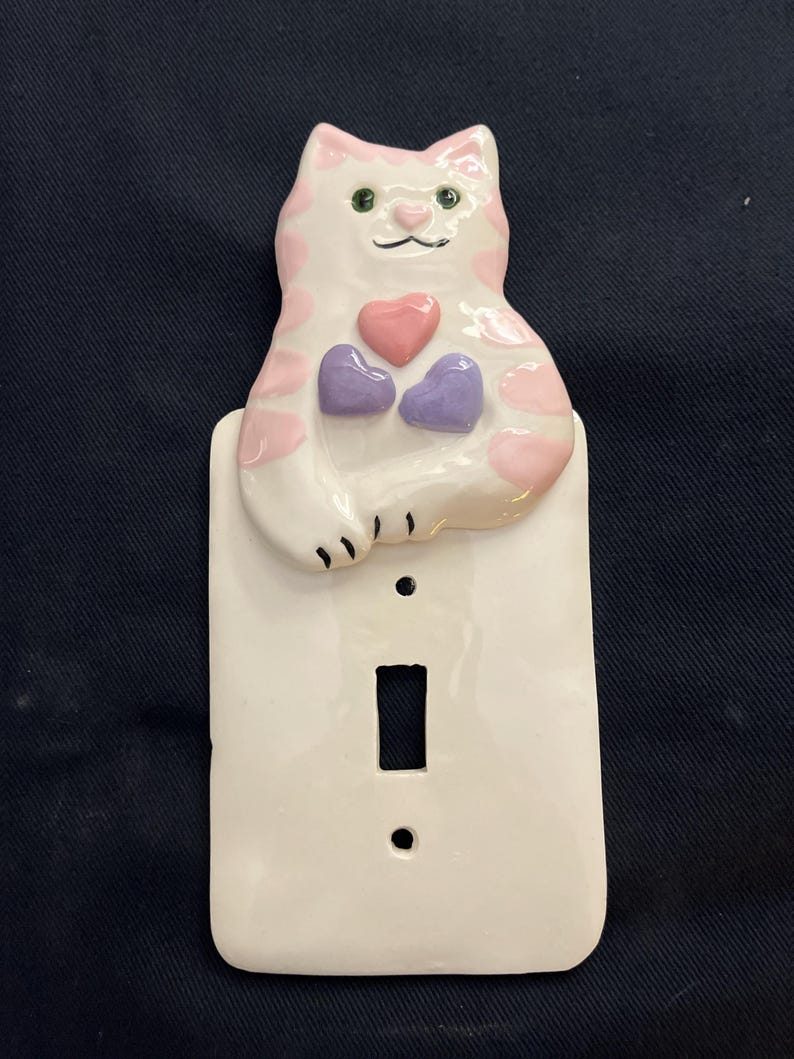 May include: A white ceramic light switch cover featuring a cat design. The cat has pink spots and holds three hearts, one pink and two purple. The switch plate is rectangular with a single switch opening and two screw holes.
