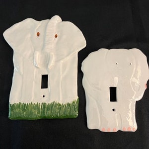May include: Two white ceramic light switch covers shaped like elephants. The elephant on the left has green grass at the bottom. The elephant on the right has pink feet.