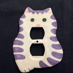 May include: A white and purple cat-shaped electrical outlet cover. The cover has a cartoon cat design with green eyes, a pink nose, and purple stripes. The cover has two openings for the electrical outlets.