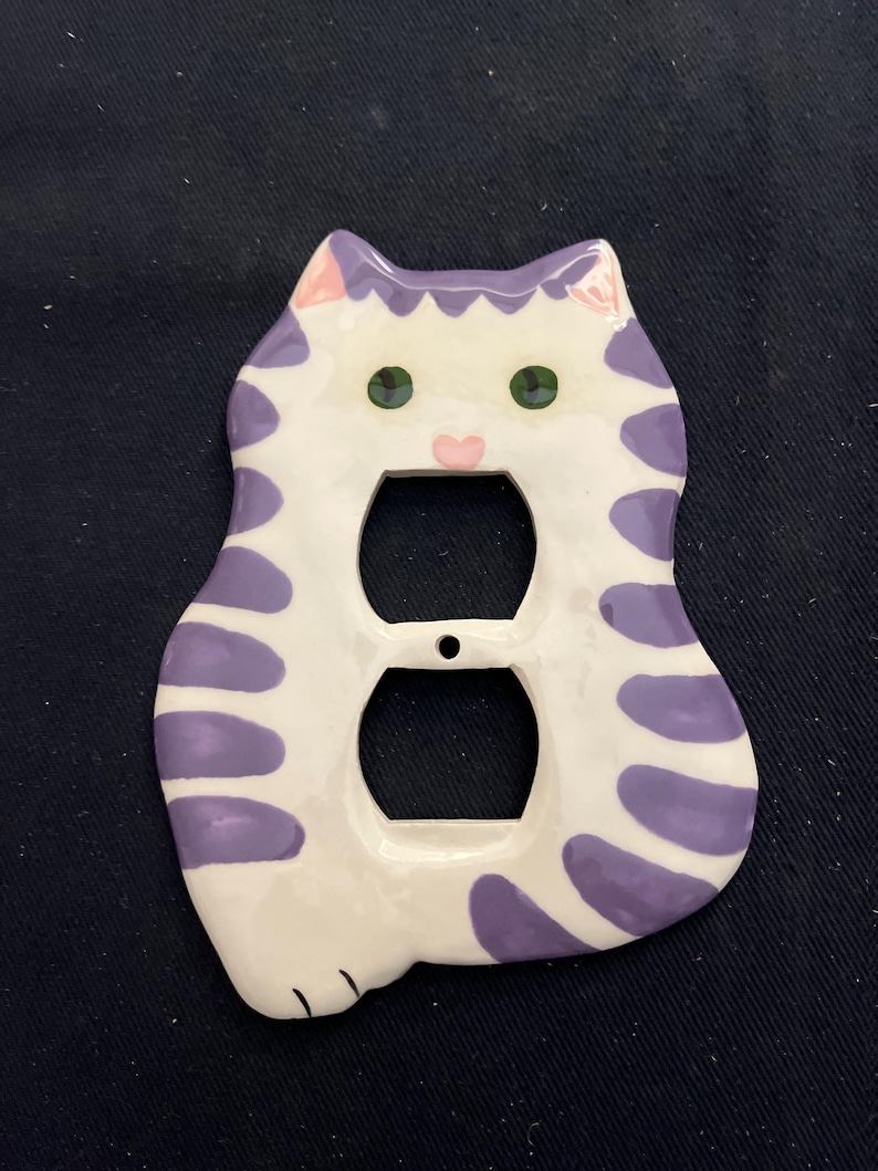 May include: A white and purple cat-shaped electrical outlet cover. The cover has a cartoon cat design with green eyes, a pink nose, and purple stripes. The cover has two openings for the electrical outlets.