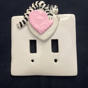 May include: White ceramic double light switch cover with a black and white striped cat resting on a pink heart. The cat has green eyes. The cover has two rectangular openings and two small circular holes.
