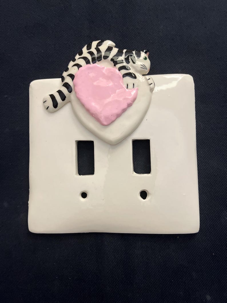 May include: White ceramic double light switch cover with a black and white striped cat resting on a pink heart. The cat has green eyes. The cover has two rectangular openings and two small circular holes.