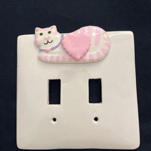 May include: White ceramic double light switch cover with a raised cat design. The cat is pink and white striped, with a pink heart and purple collar. The switch plate has two rectangular openings and two screw holes.