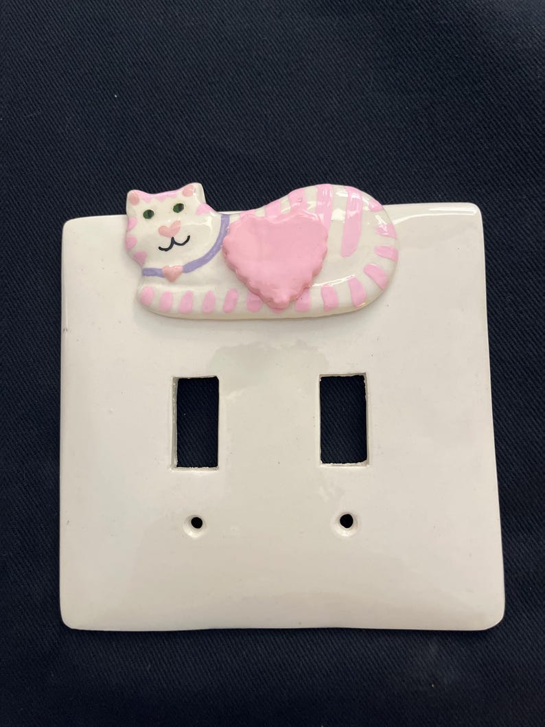May include: White ceramic double light switch cover with a raised cat design. The cat is pink and white striped, with a pink heart and purple collar. The switch plate has two rectangular openings and two screw holes.