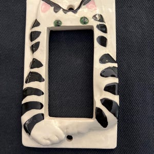 May include: A white ceramic light switch cover featuring a whimsical cat design. The cat has black stripes, green eyes, and pink ears. The cover is rectangular with a cutout for the switch and two screw holes.