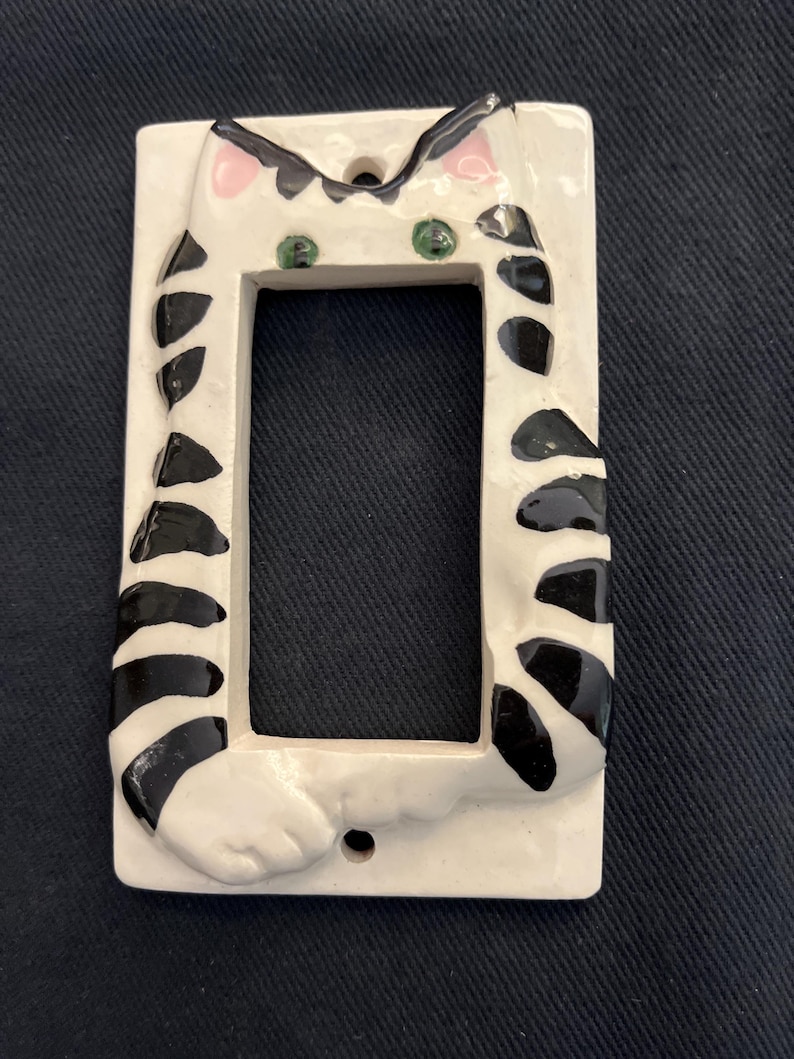 May include: A white ceramic light switch cover featuring a whimsical cat design. The cat has black stripes, green eyes, and pink ears. The cover is rectangular with a cutout for the switch and two screw holes.