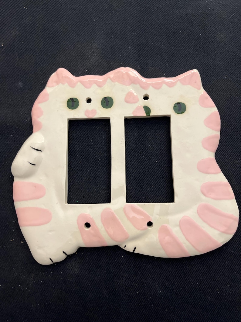 May include: A ceramic light switch cover in the shape of a cat. The cover is white with pink stripes and accents, and green eyes. It has two rectangular openings for the switches.