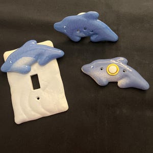 May include: Three ceramic light switch covers featuring blue dolphin designs. One cover has a white background with a light switch opening. The other two covers are individual dolphins with a white background.