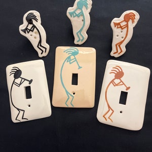May include: Five ceramic light switch covers featuring a stylized Kokopelli figure playing a flute. The covers are in various colors, including black, brown, teal, and orange.