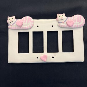May include: White ceramic four-gang light switch cover with pink and white cat figures. Each cat has a pink heart and a purple collar. The cats are lying down. The cover has a pink heart in the center.