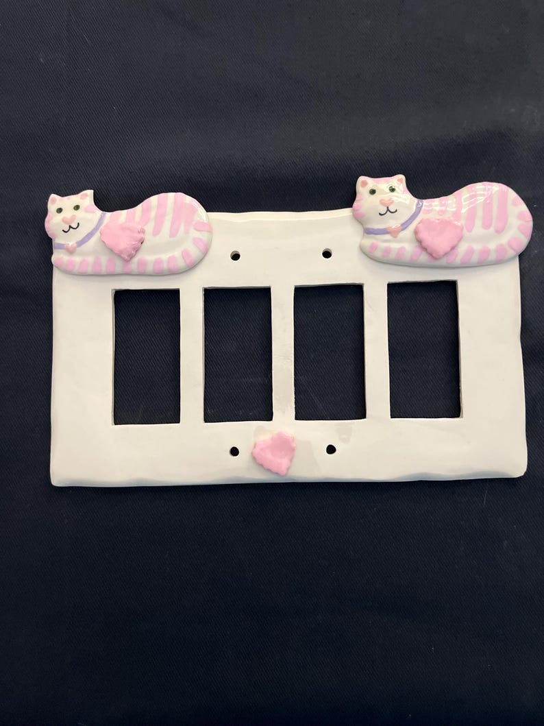 May include: White ceramic four-gang light switch cover with pink and white cat figures. Each cat has a pink heart and a purple collar. The cats are lying down. The cover has a pink heart in the center.