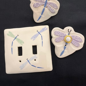 May include: A set of three white ceramic light switch covers with dragonfly designs. The covers feature blue, green, and purple dragonflies with white dots. One cover has a single switch, while the other has two switches. The single switch cover has a round, white button.