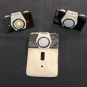 May include: Three ceramic light switch covers shaped like vintage cameras. The top two covers are black and white with the word "FLASH" printed on them. The bottom cover is white with a black and white camera design and the word "SMILE" printed on it.