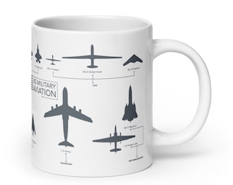 Puede incluir: Una taza de cerámica blanca con un gráfico en blanco y negro de siluetas de varios aviones militares estadounidenses. El gráfico se titula "US Military Aviation" e incluye los nombres de los aviones, como "F-22 Raptor", "B-2 Spirit" y "C-5 Galaxy".