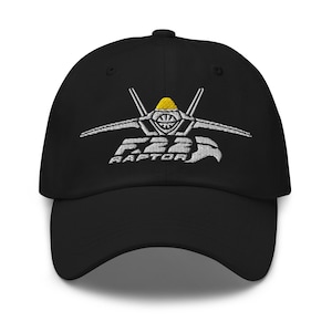 May include: Black baseball cap with an embroidered F-22 Raptor jet fighter design in white and yellow.