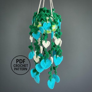 May include: A handmade crochet hanging planter with a heart-shaped leaf design. The planter features a green top, with trailing vines of blue and white heart-shaped leaves. The text "PDF CROCHET PATTERN" is in a circle at the bottom.