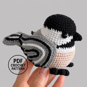 May include: A crocheted chickadee bird figurine featuring a black head, white cheeks, and a grey and black striped wing. The body has a light beige underside and small black feet. A circular black and white logo reads PDF CROCHET PATTERN.