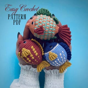 Pode incluir: Três brinquedos de peixe de crochê em diferentes cores. Os peixes são vermelho, azul e laranja com barbatanas amarelas. O texto "Easy Crochet Pattern PDF" é visível na imagem.