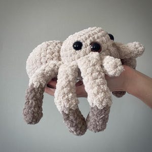 May include: A plush, crocheted spider toy in shades of cream and gray. The spider has large, black and brown eyes and eight textured legs. The toy is soft and cuddly, perfect for children or as a decorative item.
