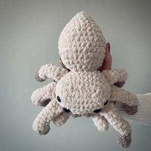 May include: A plush, cream-colored spider toy with eight legs and a rounded body. The spider's legs and body are made of a soft, textured material. The spider has small, black eyes and gray accents on the tips of its legs.
