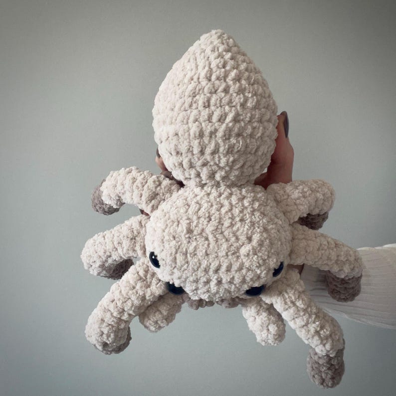 May include: A plush, cream-colored spider toy with eight legs and a rounded body. The spider's legs and body are made of a soft, textured material. The spider has small, black eyes and gray accents on the tips of its legs.