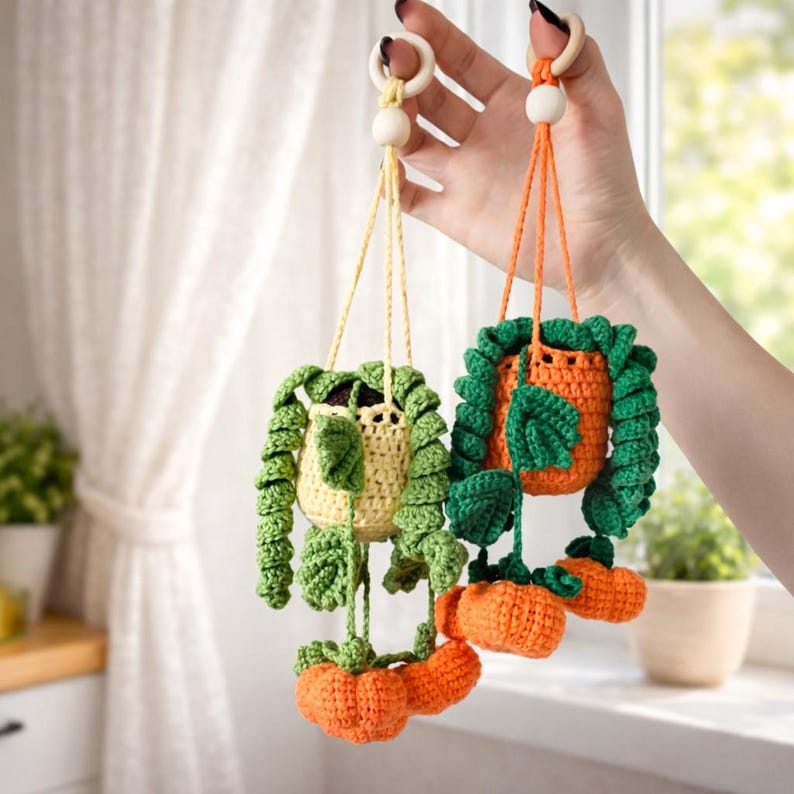 May include: Two hanging crochet plant decorations featuring small pots in yellow and orange. Each pot has green crocheted vines with leaves and small orange pumpkin-shaped accents. The items are suspended by yarn cords attached to wooden rings.