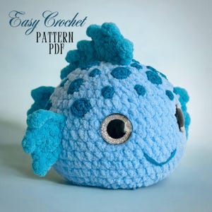 Puede incluir: Un pez de crochet azul y turquesa con un ojo blanco y negro. El pez tiene una aleta puntiaguda en la parte superior de la cabeza y una aleta pequeña a cada lado. El pez es un juguete de peluche.