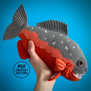 Op de afbeelding: Een gehaakt vis speelgoed in grijstinten en oranje, met witte accenten. De vis heeft een gedetailleerde staart, vinnen en een lachende mond met tanden. De tekst "PDF CROCHET PATTERN" is zichtbaar. Het speelgoed wordt door een hand vastgehouden.