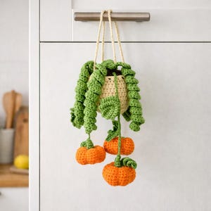 May include: A crocheted hanging plant featuring a cream-colored pot, long green curly vines, and three small orange pumpkins. The plant is suspended by cream-colored yarn strings, showcasing detailed stitch work on the leaves and fruit.