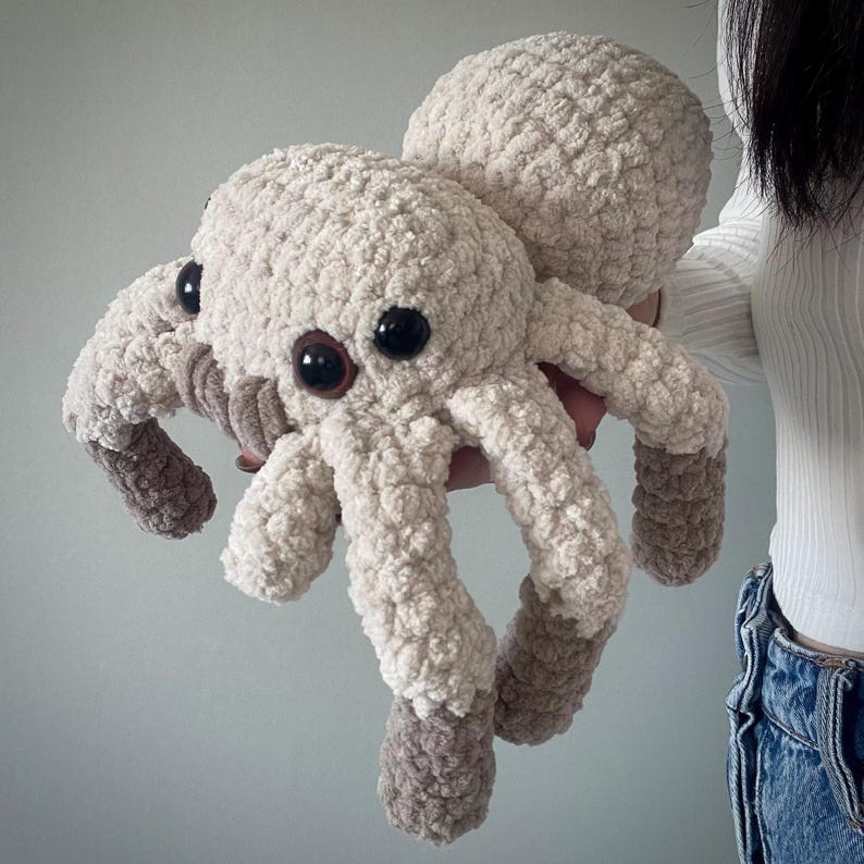 May include: A plush, beige spider toy with large black eyes and brown accents on its legs. The soft, crocheted spider has a rounded body and eight legs, perfect for a child's toy or a unique home decor item.