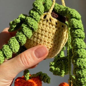 May include: A crocheted hanging plant in a light yellow pot with dark brown soil, featuring long, trailing green vines with textured leaves. Below the pot, a small orange crocheted pumpkin with green leaves is visible, all crafted from yarn.